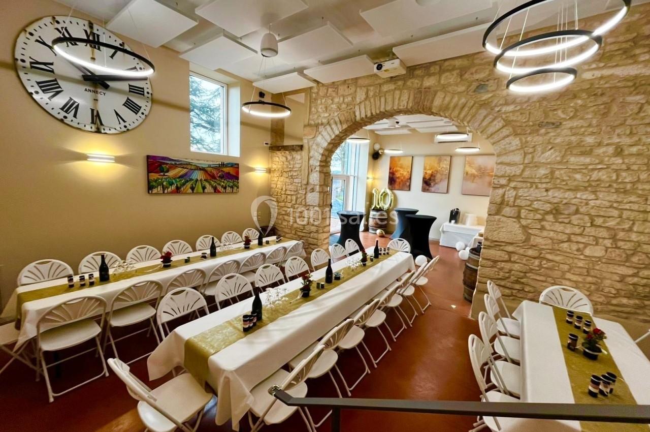 Salle décorée avec des tables alignées pour un événement, murs en pierre, grande horloge et éclairage moderne.