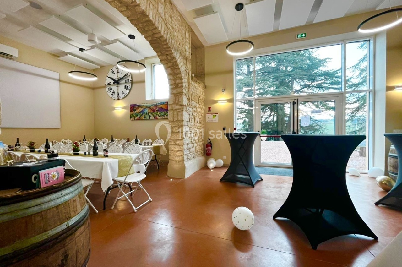 Salle de réception lumineuse avec tables dressées, tonneau décoratif, grandes baies vitrées et vue sur des arbres.
