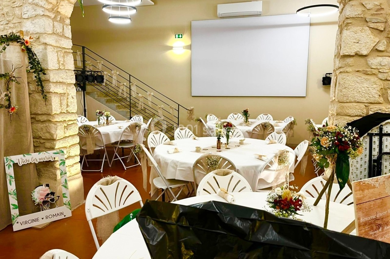 Salle de réception décorée avec des tables rondes, nappes blanches, chaises pliantes et arrangements floraux.