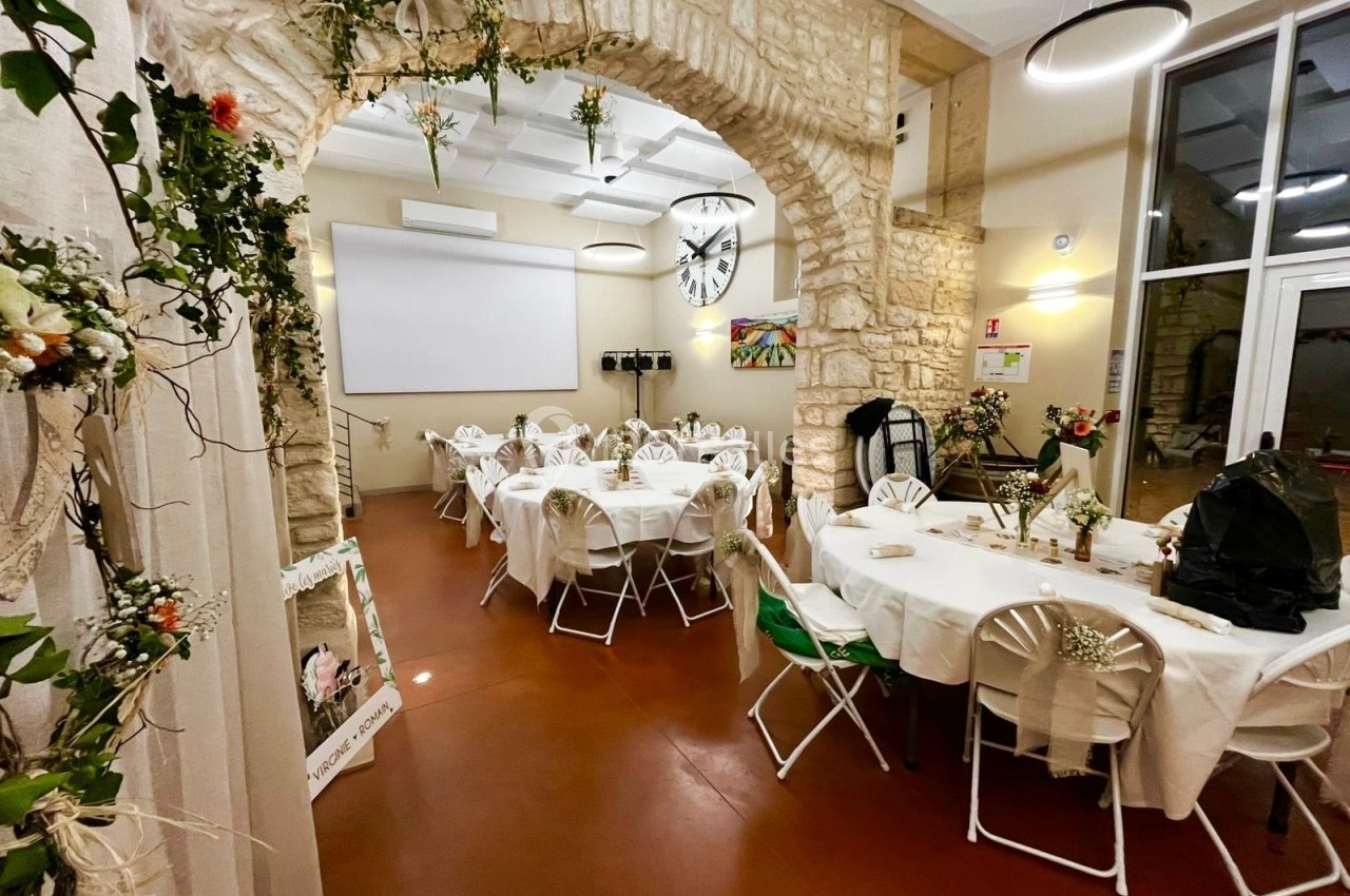 Salle décorée pour un événement, avec tables rondes dressées, fleurs et arches en pierre apparente.