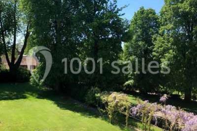 Glycine en fleurs grimpant sur une pergola, avec une maison en briques et des arbustes en arrière-plan.