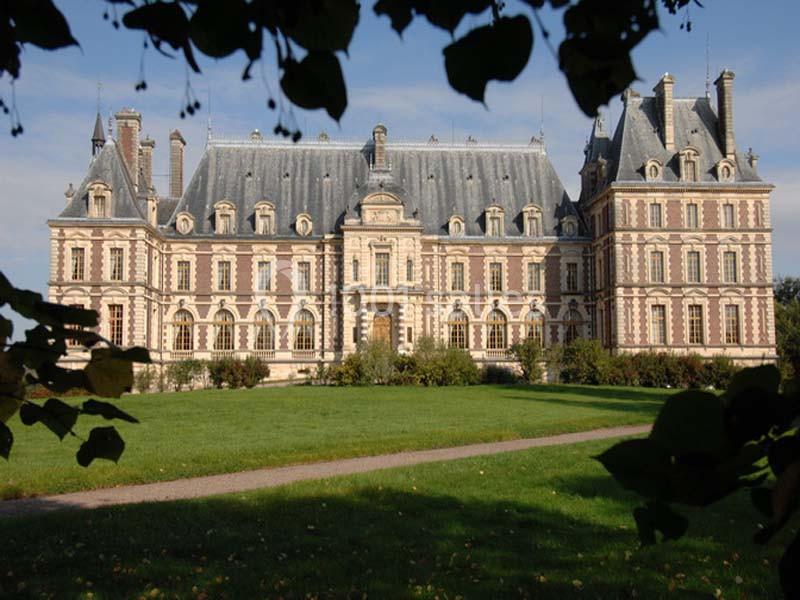 Façade d'un château historique en pierre et brique, entouré d'un parc verdoyant sous un ciel dégagé.