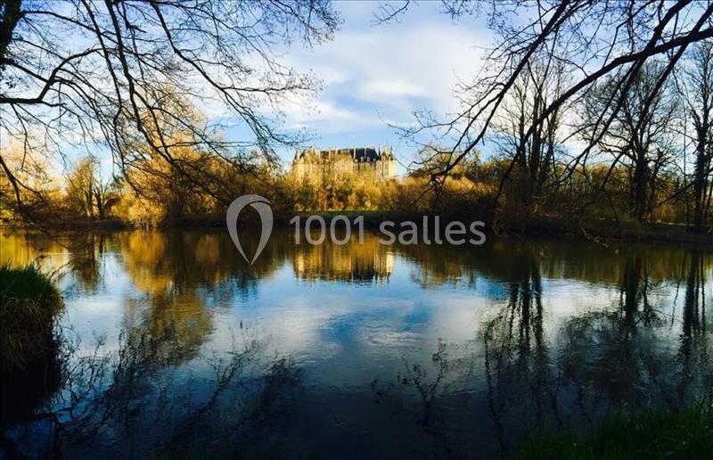 Vue d'un château au loin, entouré d'arbres, reflété dans un étang calme sous un ciel partiellement nuageux.