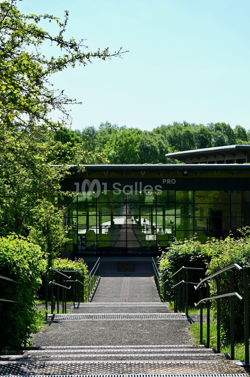 Escalier bordé de verdure menant à un bâtiment moderne avec une façade vitrée reflétant un paysage arboré.