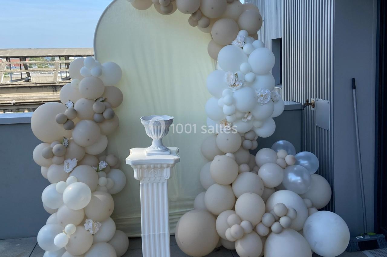 Arche décorative en ballons beige et blanc avec un piédestal blanc sur une terrasse extérieure.