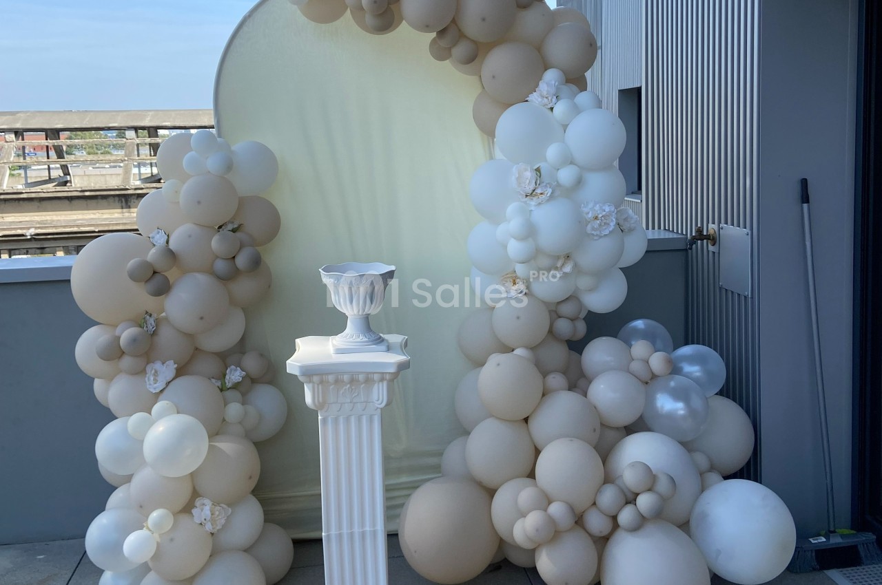Arche décorative en ballons beige et blanc avec un piédestal blanc sur une terrasse extérieure.