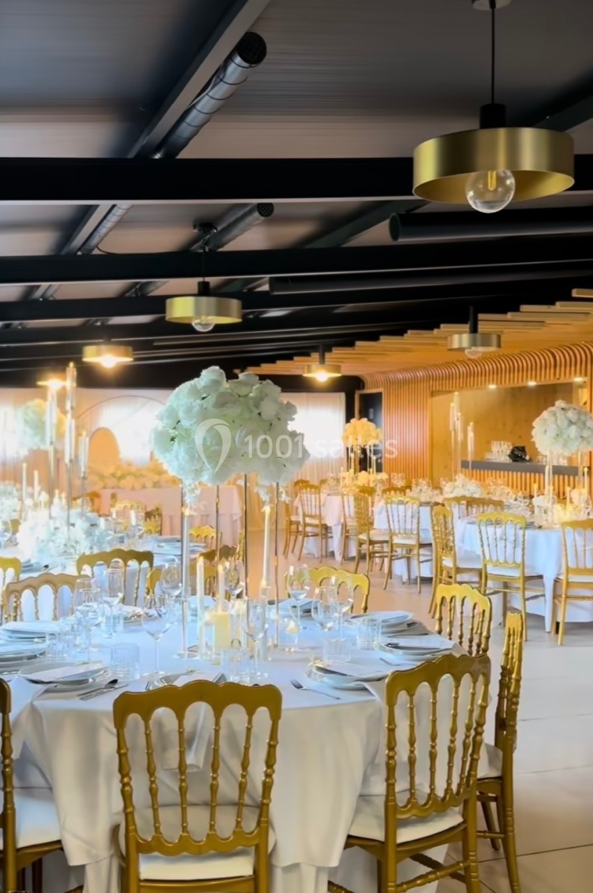Salle de réception élégante avec tables rondes décorées de nappes blanches, chaises dorées et centres de table fleuris.