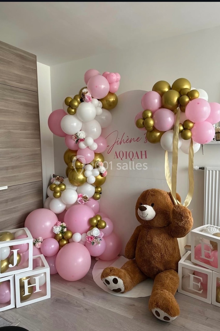 Décoration festive avec ballons roses, blancs et dorés, grand ours en peluche et cubes décoratifs dans une pièce lumineuse.