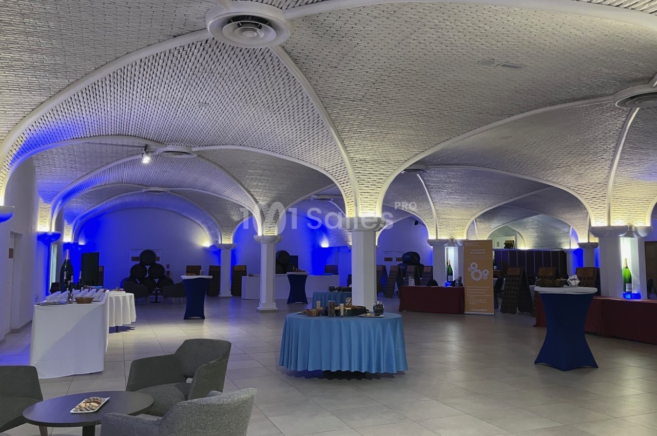 Salle voûtée éclairée en bleu avec tables dressées, fauteuils et stands d'exposition installés.