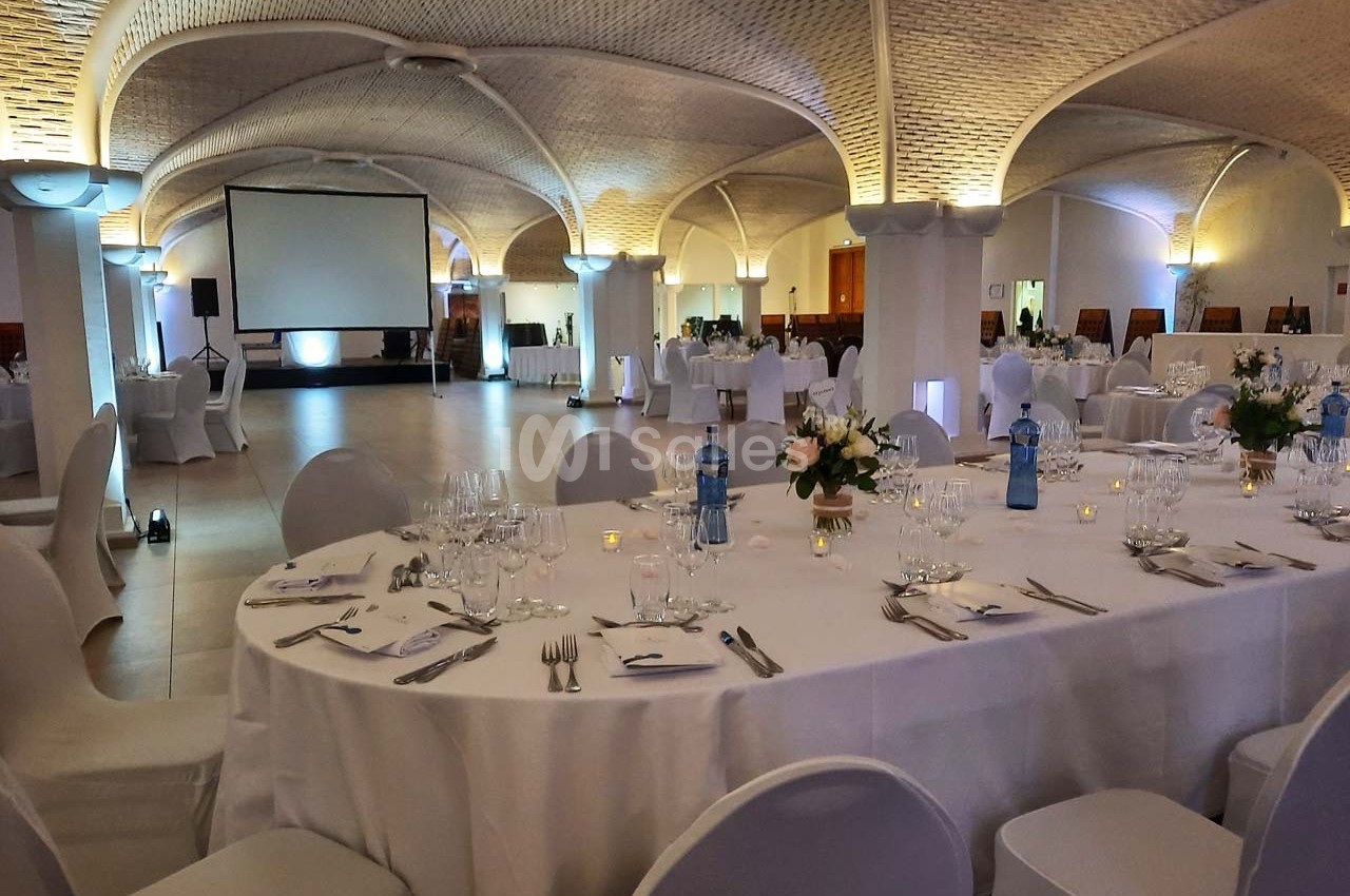 Salle de réception élégante avec tables rondes dressées, décor floral et éclairage tamisé sous un plafond voûté.