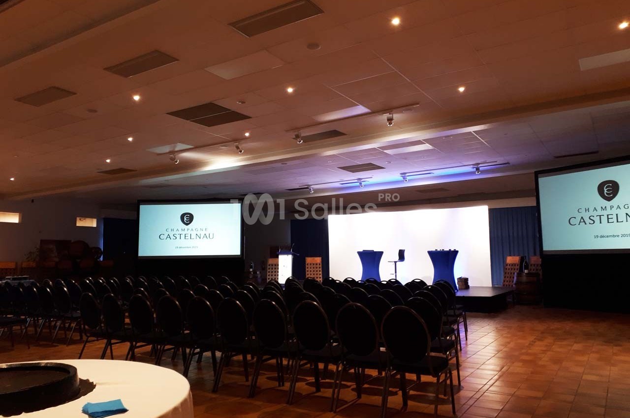Salle de conférence avec rangées de chaises, écrans de projection allumés et estrade éclairée.