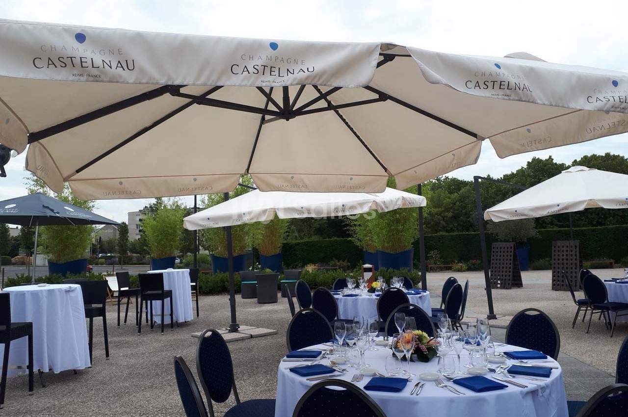 Tables dressées avec nappes blanches et parasols blancs sur une terrasse extérieure, entourée de verdure.