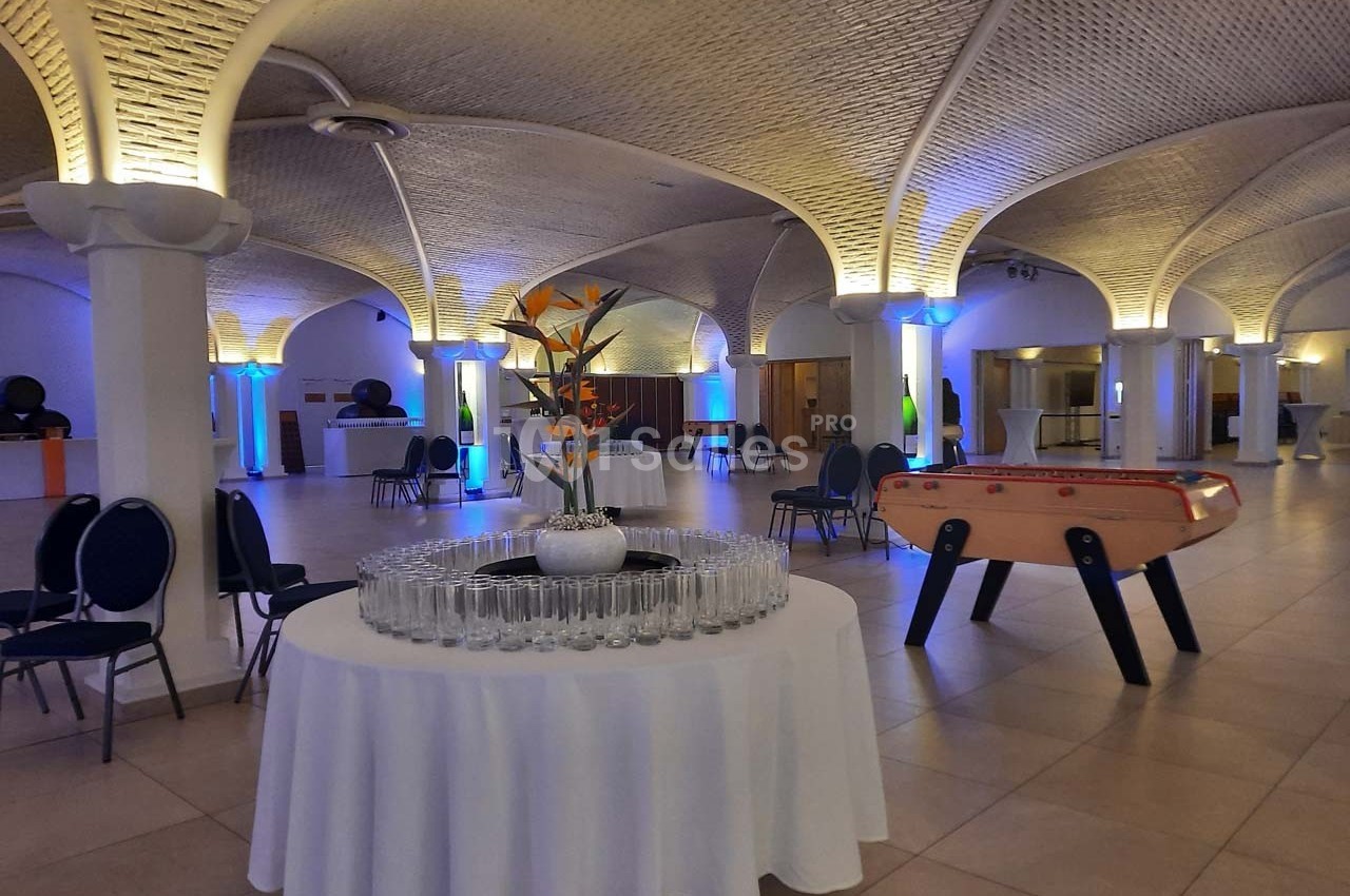 Salle voûtée avec tables dressées, chaises, baby-foot et éclairage d'ambiance coloré.