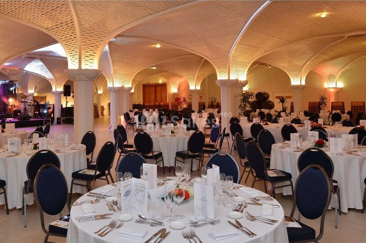 Salle de réception élégante avec tables rondes dressées, nappes blanches et éclairage chaleureux sous un plafond voûté.