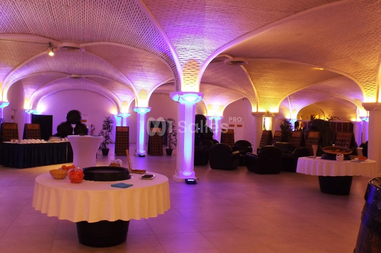 Salle voûtée éclairée avec des lumières colorées, tables rondes dressées et fauteuils disposés en arrière-plan.