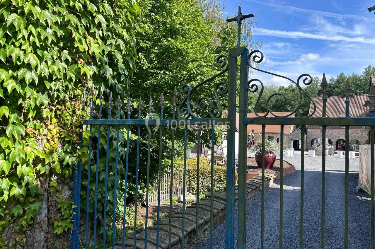 Portail en fer forgé ouvrant sur une cour pavée, entouré de végétation et de bâtiments en arrière-plan.