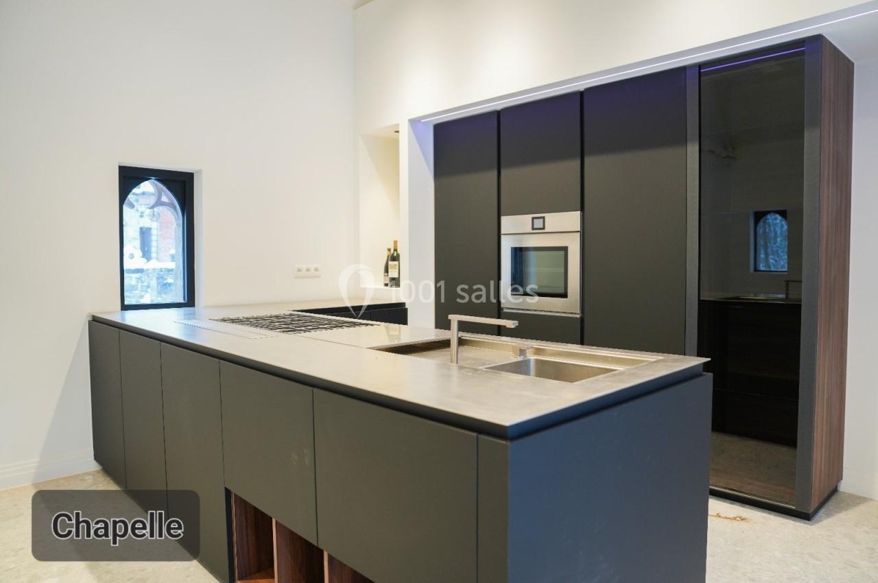 Cuisine moderne avec îlot central en acier inoxydable, placards noirs et fenêtres en ogive laissant entrer la lumière.