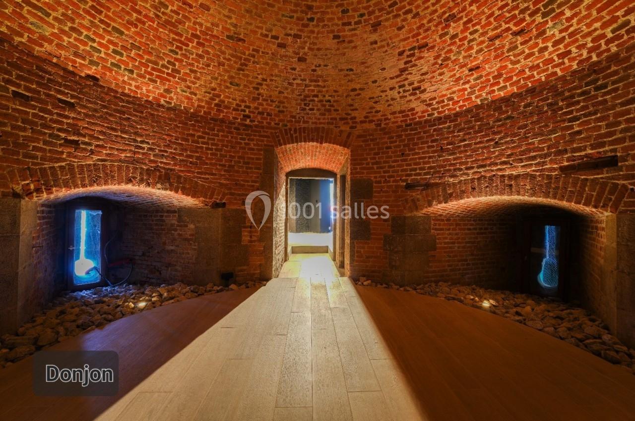 Salle voûtée en briques rouges avec des ouvertures illuminées et un sol en bois éclairé par une lumière centrale.