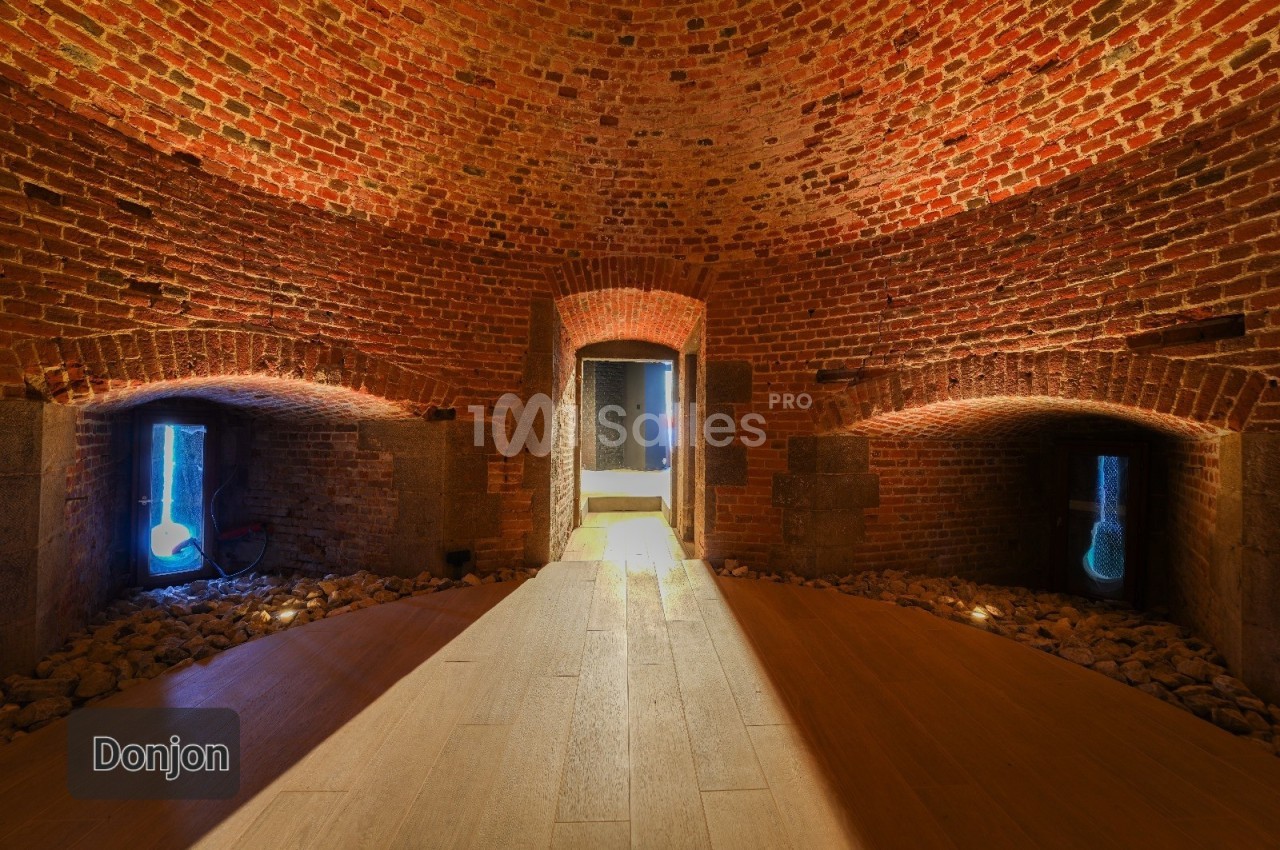 Salle voûtée en briques rouges avec des ouvertures illuminées et un sol en bois éclairé par une lumière centrale.