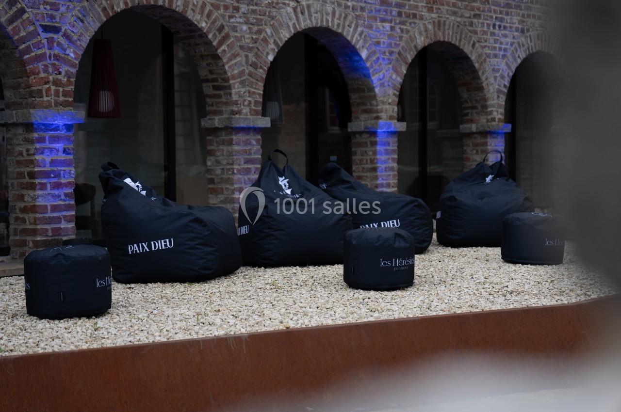 Poufs noirs marqués ’Paix Dieu’ et ’les Héritiers’ disposés sur un sol de gravier devant un bâtiment en briques.