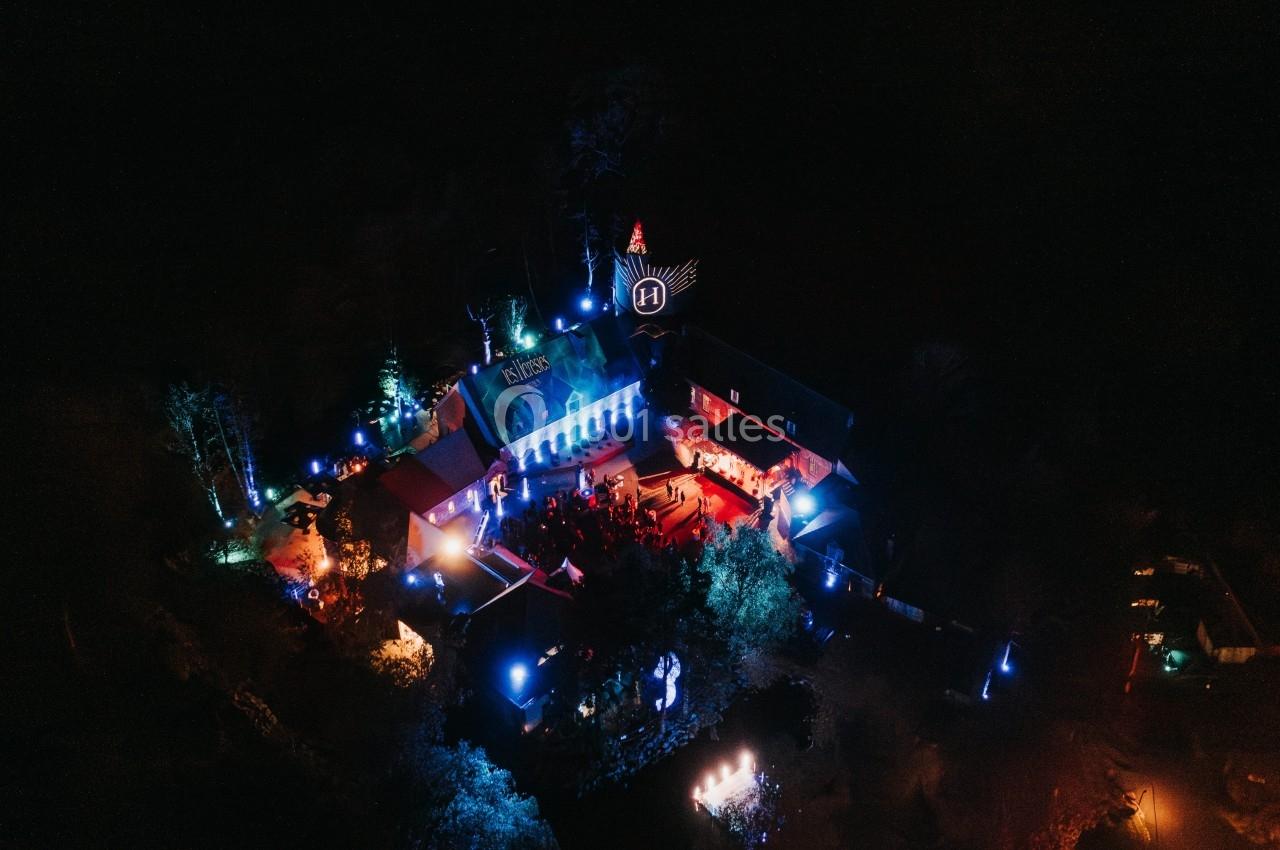 Vue aérienne nocturne d'un bâtiment illuminé par des lumières colorées, entouré de végétation sombre.