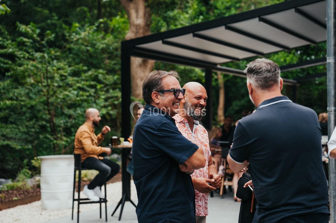 Trois hommes discutent debout dans un espace extérieur avec des arbres et des personnes en arrière-plan.