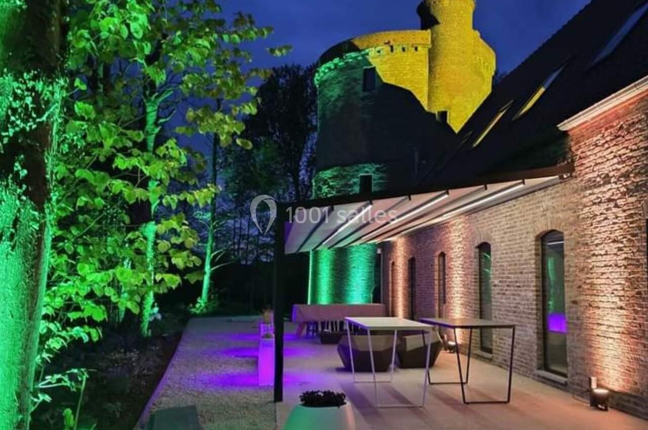 Terrasse moderne éclairée en soirée avec des lumières colorées, près d'un bâtiment en pierre et une tour médiévale.
