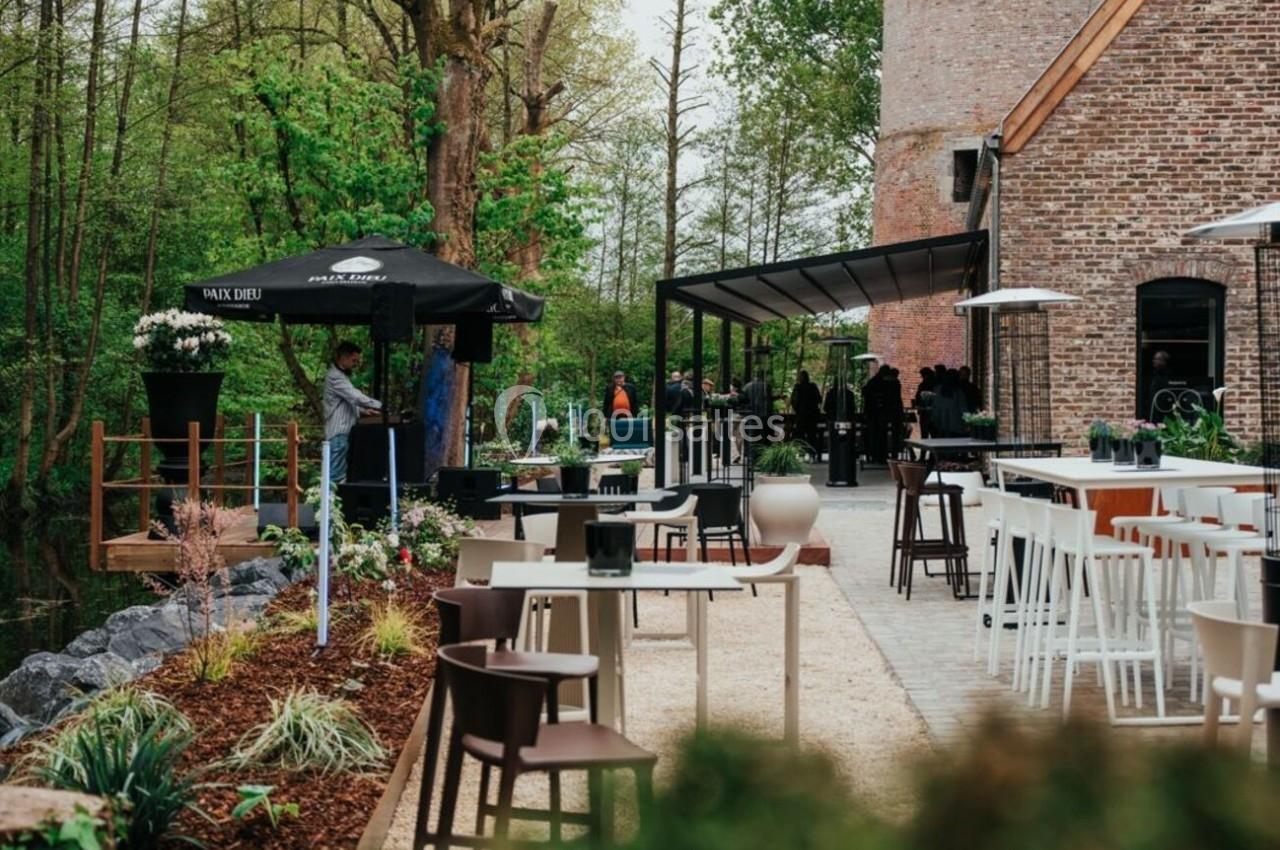 Terrasse extérieure d'un restaurant avec mobilier moderne, entourée de verdure et d'un bâtiment en briques avec une tour.