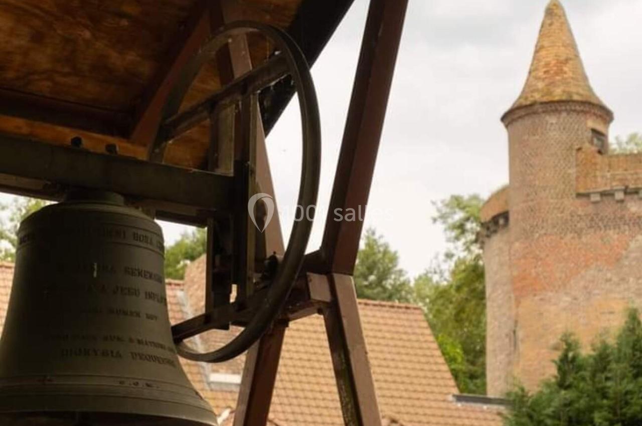 Cloche suspendue sous un toit en bois, avec une tour en briques et des toits en arrière-plan.