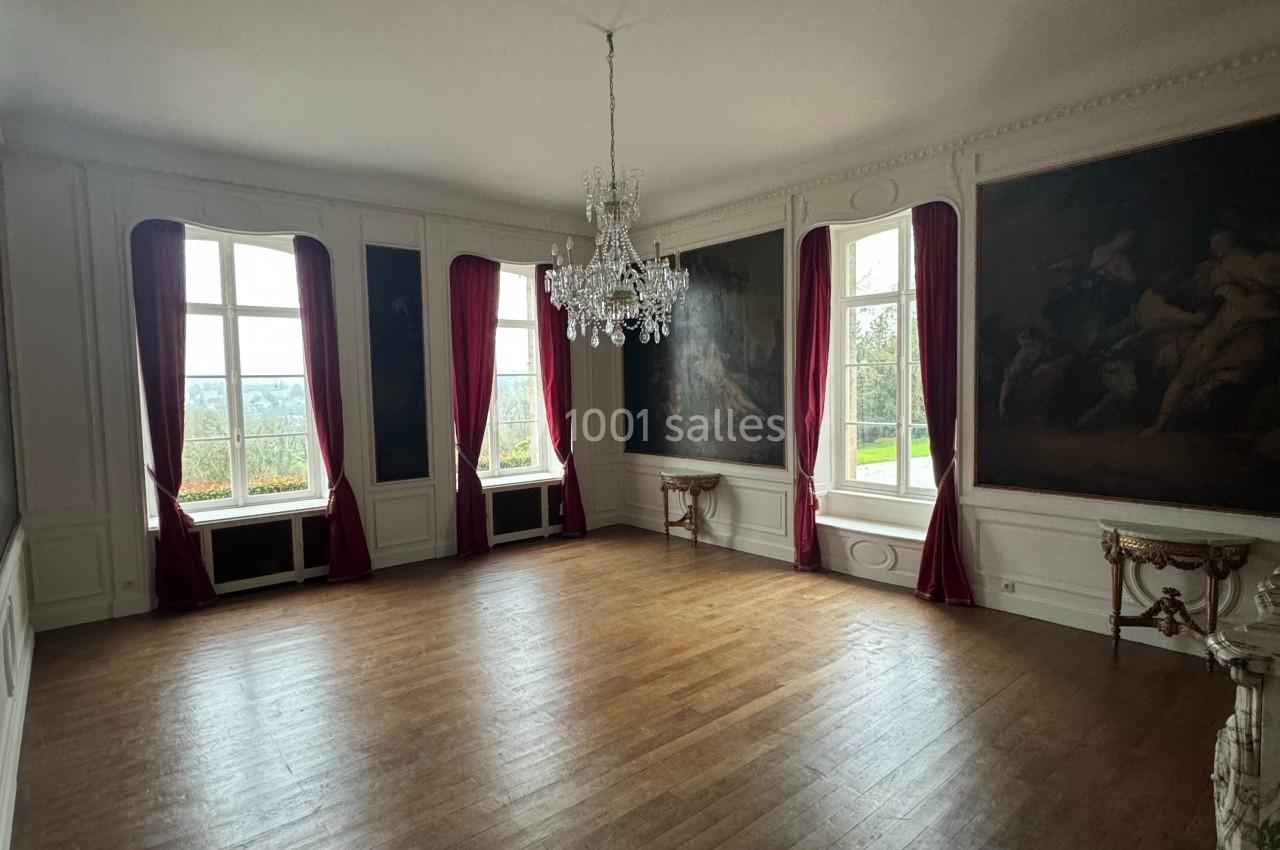 Salle lumineuse avec parquet en bois, murs ornés de tableaux, rideaux rouges et un lustre central.