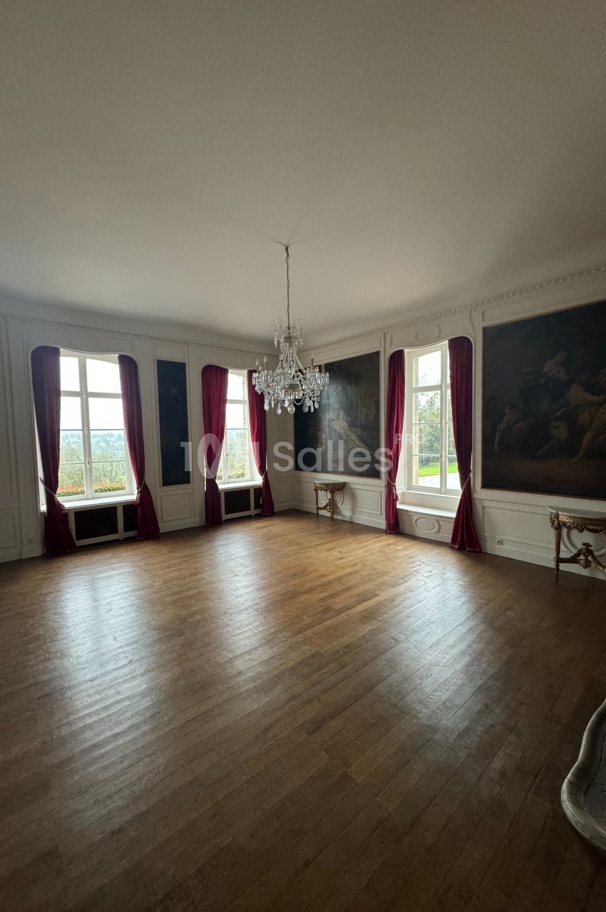 Salle lumineuse avec parquet en bois, murs ornés de tableaux, rideaux rouges et un lustre central.