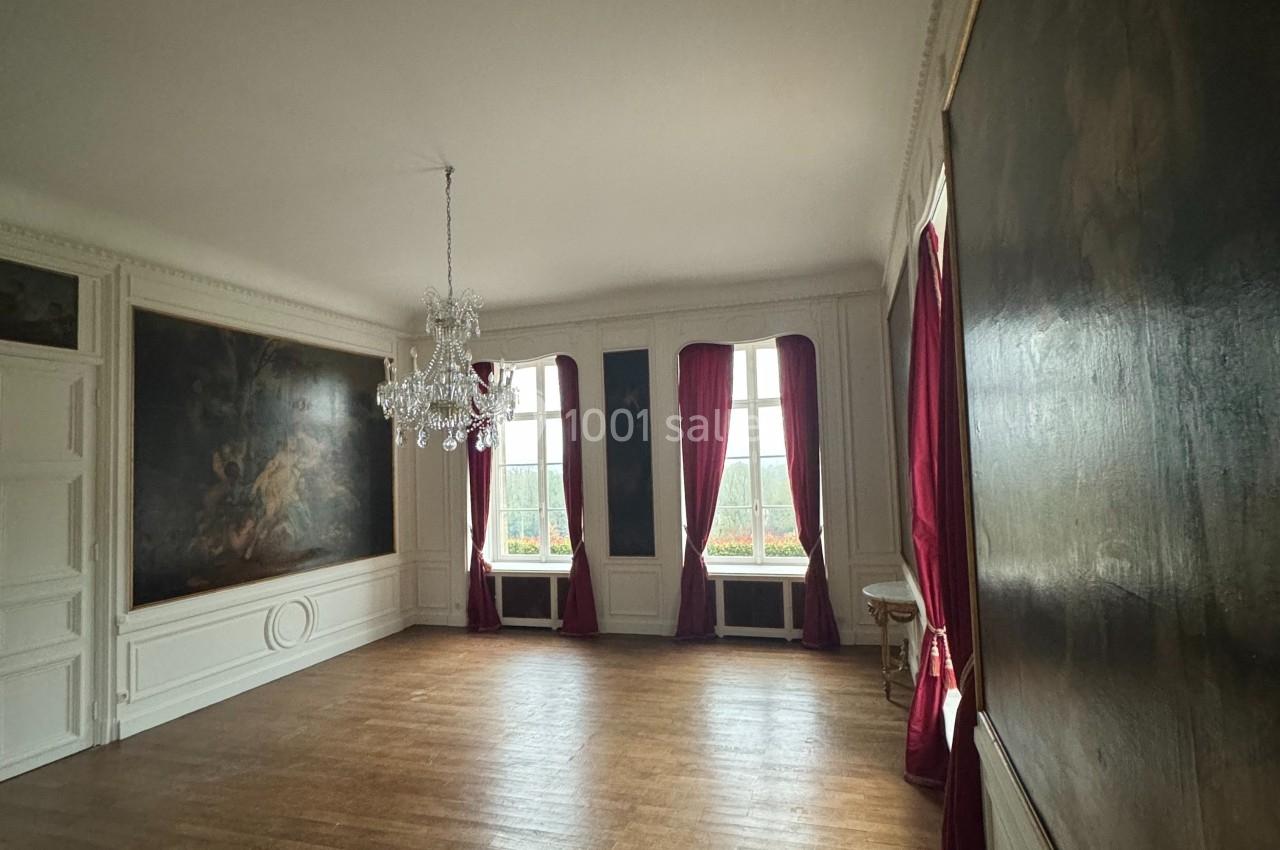 Salle lumineuse avec parquet en bois, murs ornés de tableaux, rideaux rouges et lustre suspendu au plafond.