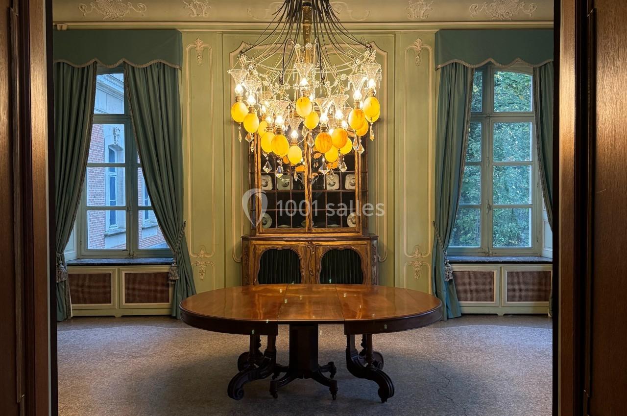Salle élégante avec un lustre imposant, table ronde en bois, chaises vertes et grandes fenêtres encadrées de rideaux.