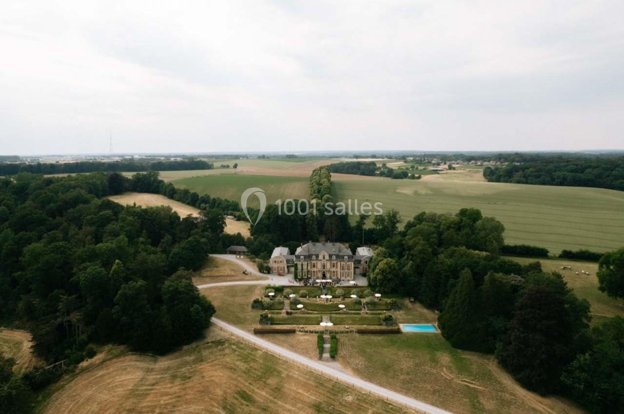 Vue aérienne d'un château entouré de jardins, de champs et de forêts sous un ciel légèrement nuageux.