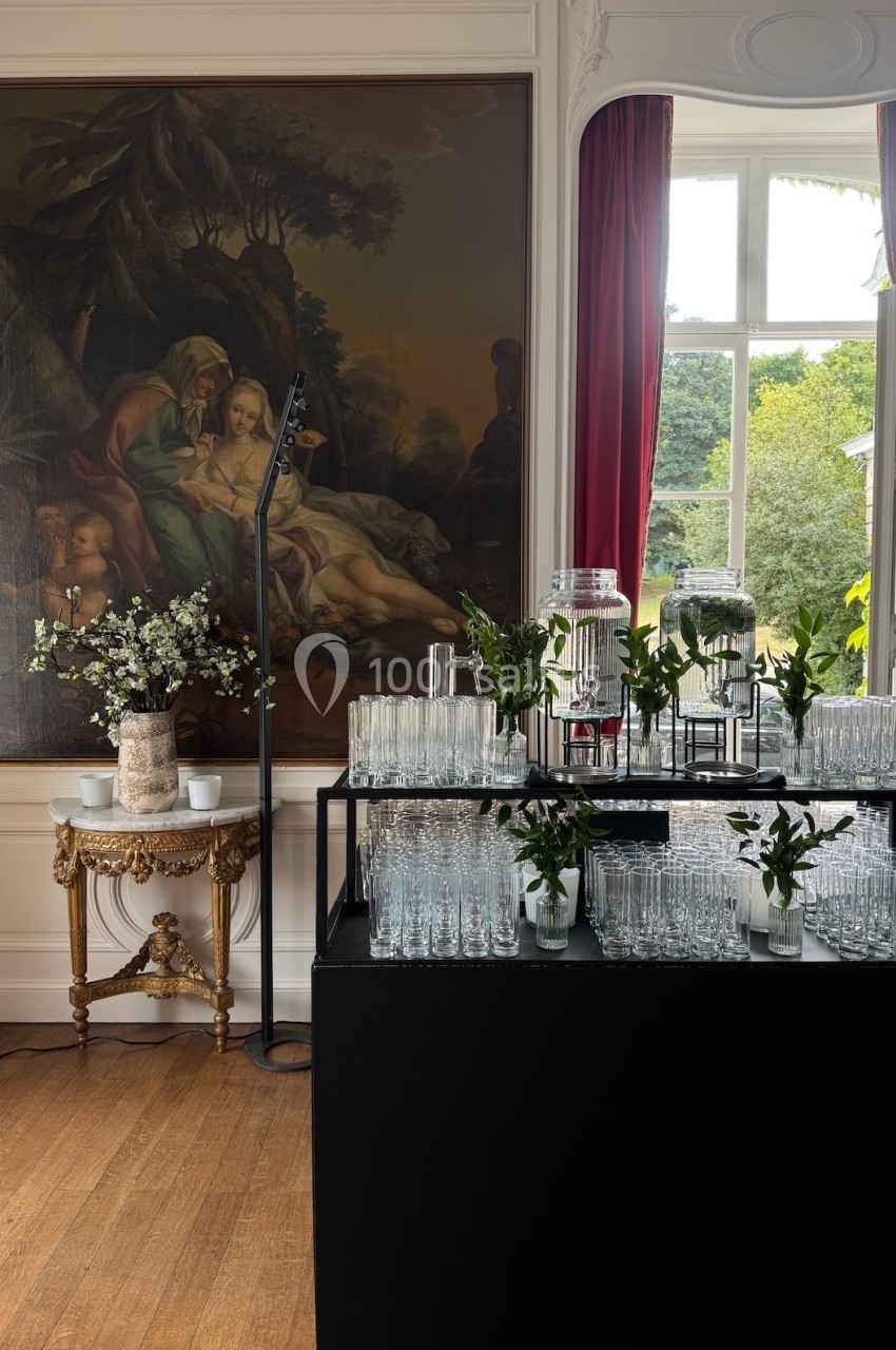 Table avec verres et décorations florales dans une pièce élégante, devant une grande peinture et une fenêtre lumineuse.