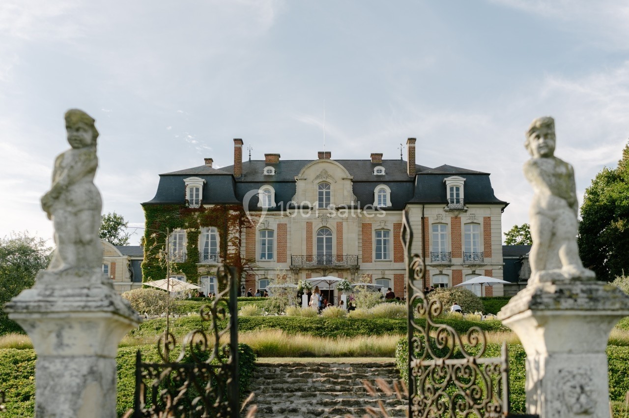 Façade d'un château historique entouré de jardins, vue depuis une grille en fer forgé ornée de statues.