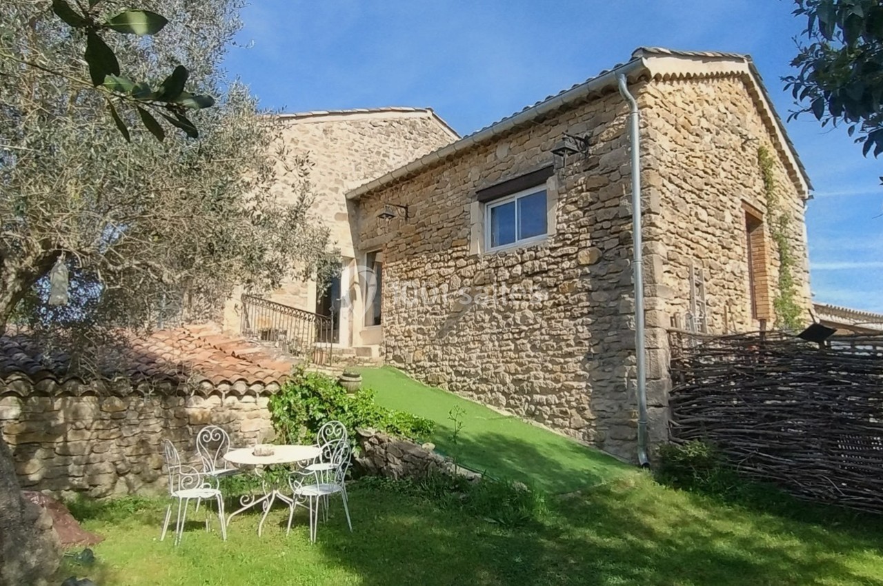 Maison en pierre avec jardin, table et chaises blanches, entourée d'arbres et d'une clôture en bois tressé.