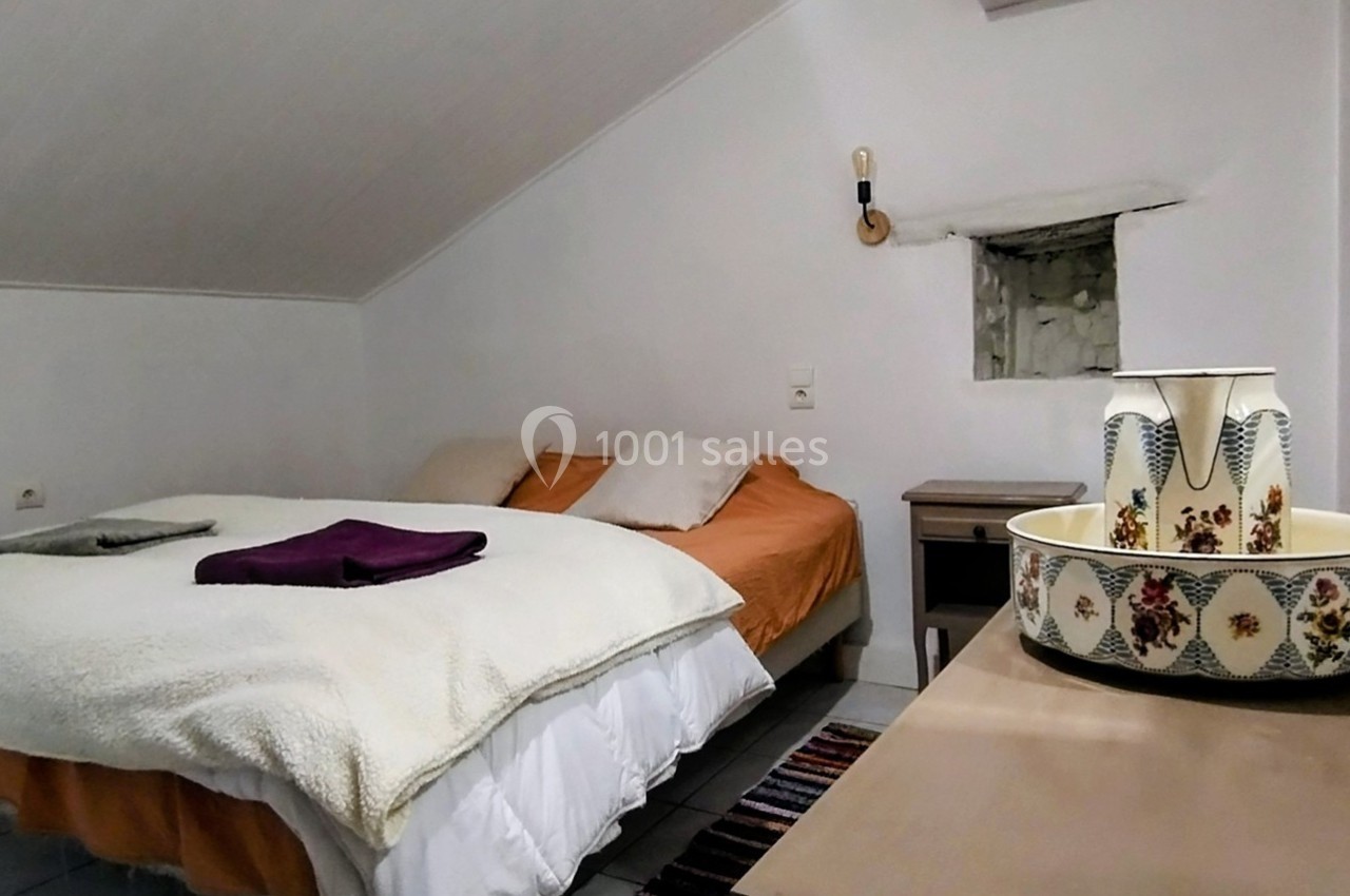 Chambre mansardée avec lit double, table de chevet, miroir et décoration en céramique sur une commode en bois.