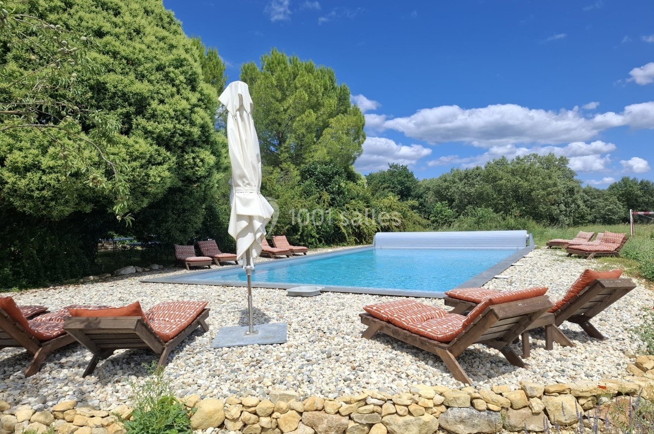 Piscine extérieure entourée de transats en bois avec coussins rouges, située dans un jardin arboré sous un ciel bleu.