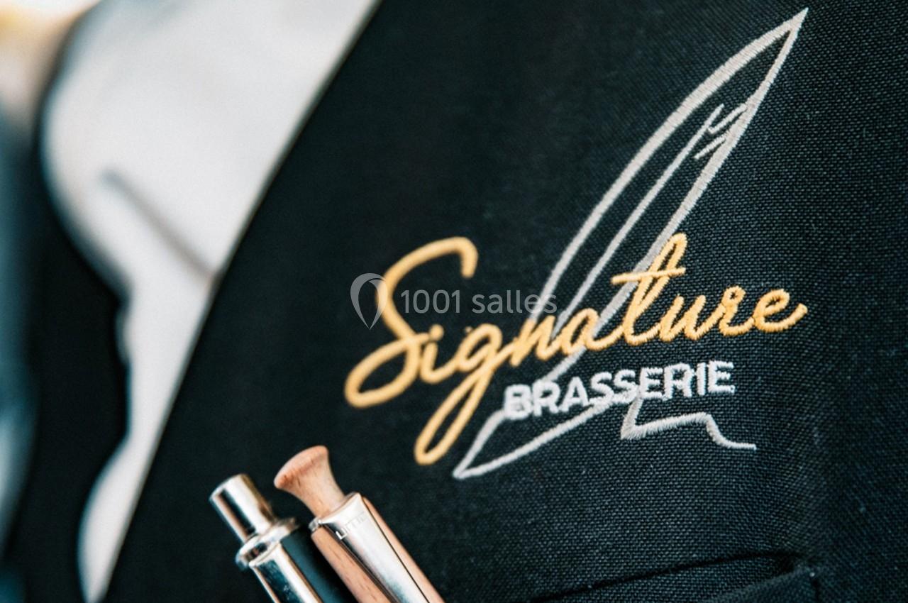 Logo ’Signature Brasserie’ brodé sur un tissu noir, avec des stylos insérés dans une poche.