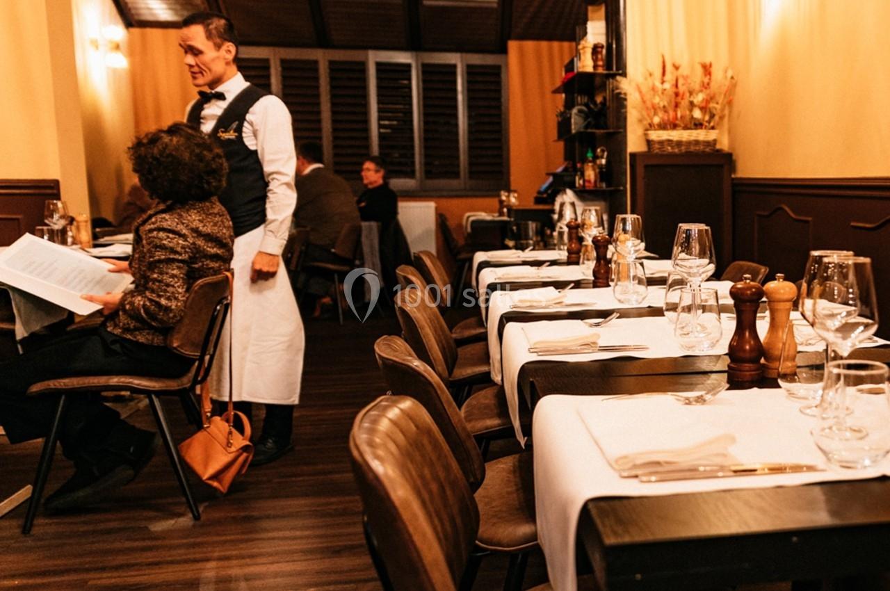 Salle de restaurant chaleureux avec des tables dressées, un serveur en uniforme et des clients assis.