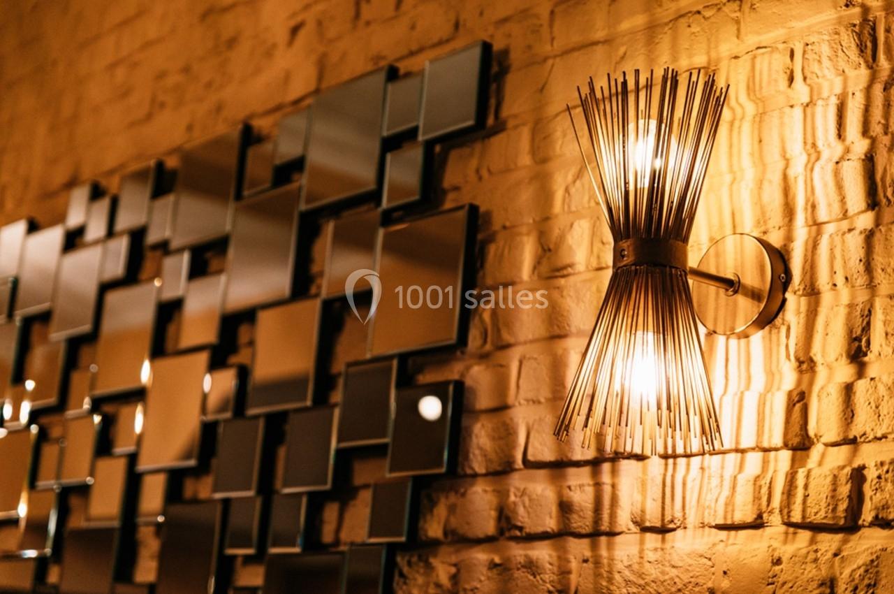 Applique murale dorée allumée sur un mur en briques, à côté d'un assemblage de miroirs géométriques.