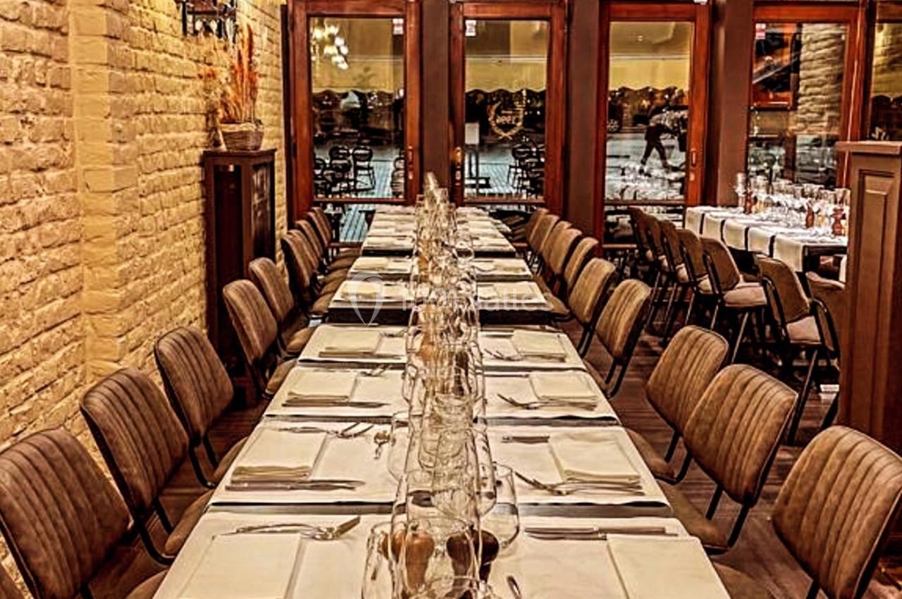 Salle de restaurant avec deux longues tables dressées, entourées de chaises, et une vue sur l'extérieur à travers des baies…