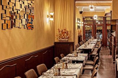 Salle de restaurant avec tables dressées, banquettes en bois, bar en bois et étagères garnies de verres et bouteilles.