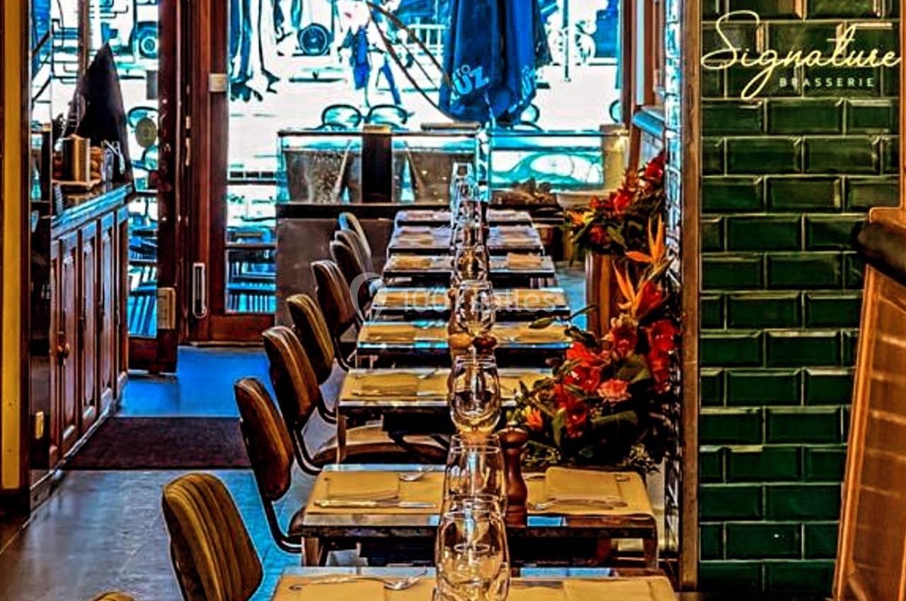 Salle de restaurant avec une longue table dressée, chaises alignées et décoration florale près d'un mur en carreaux verts.