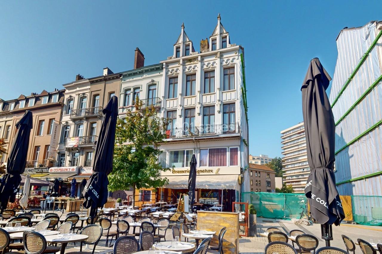 Terrasse de café avec tables et chaises vides devant des bâtiments anciens sous un ciel dégagé.