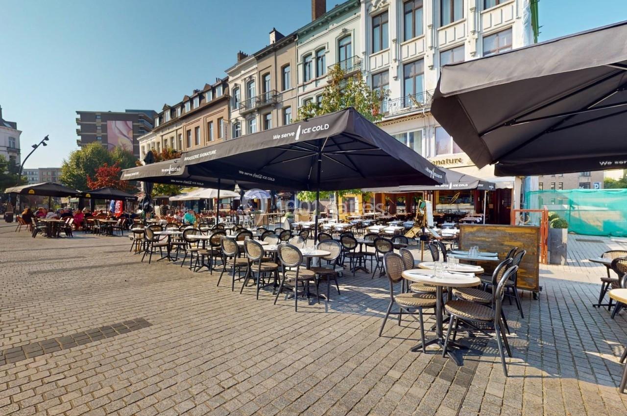 Terrasse de café avec tables et chaises disposées sur une place pavée, entourée de bâtiments urbains.