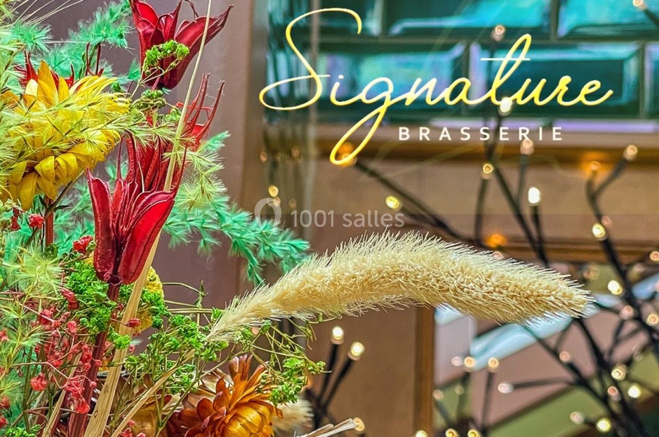 Composition florale colorée avec des fleurs séchées et feuillages devant un panneau lumineux indiquant ’Signature Brasserie’.