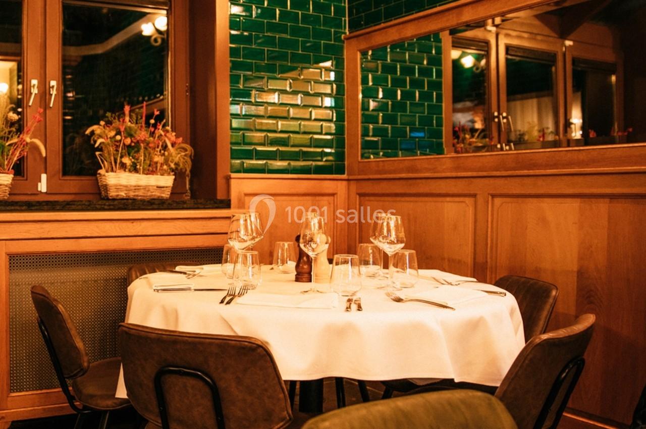 Salle de restaurant avec table ronde dressée, chaises en cuir, murs en bois et carreaux verts brillants.
