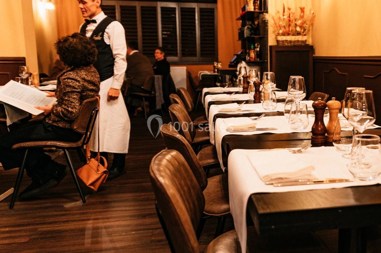 Salle de restaurant chaleureuse avec tables dressées, un serveur interagit avec une cliente assise.